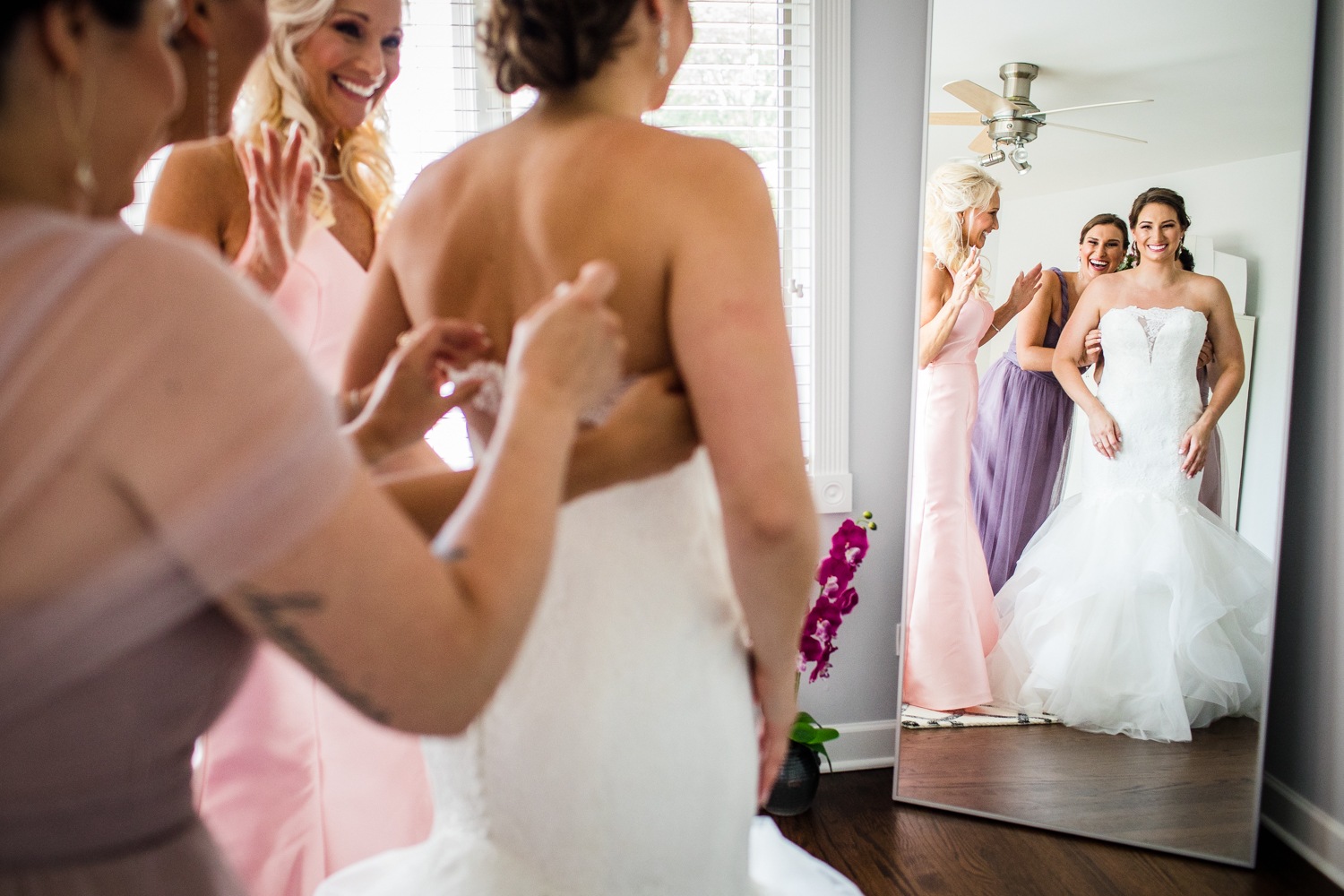 A bride laughs with her wedding party reflected in a mirror before her Glen Club wedding. 