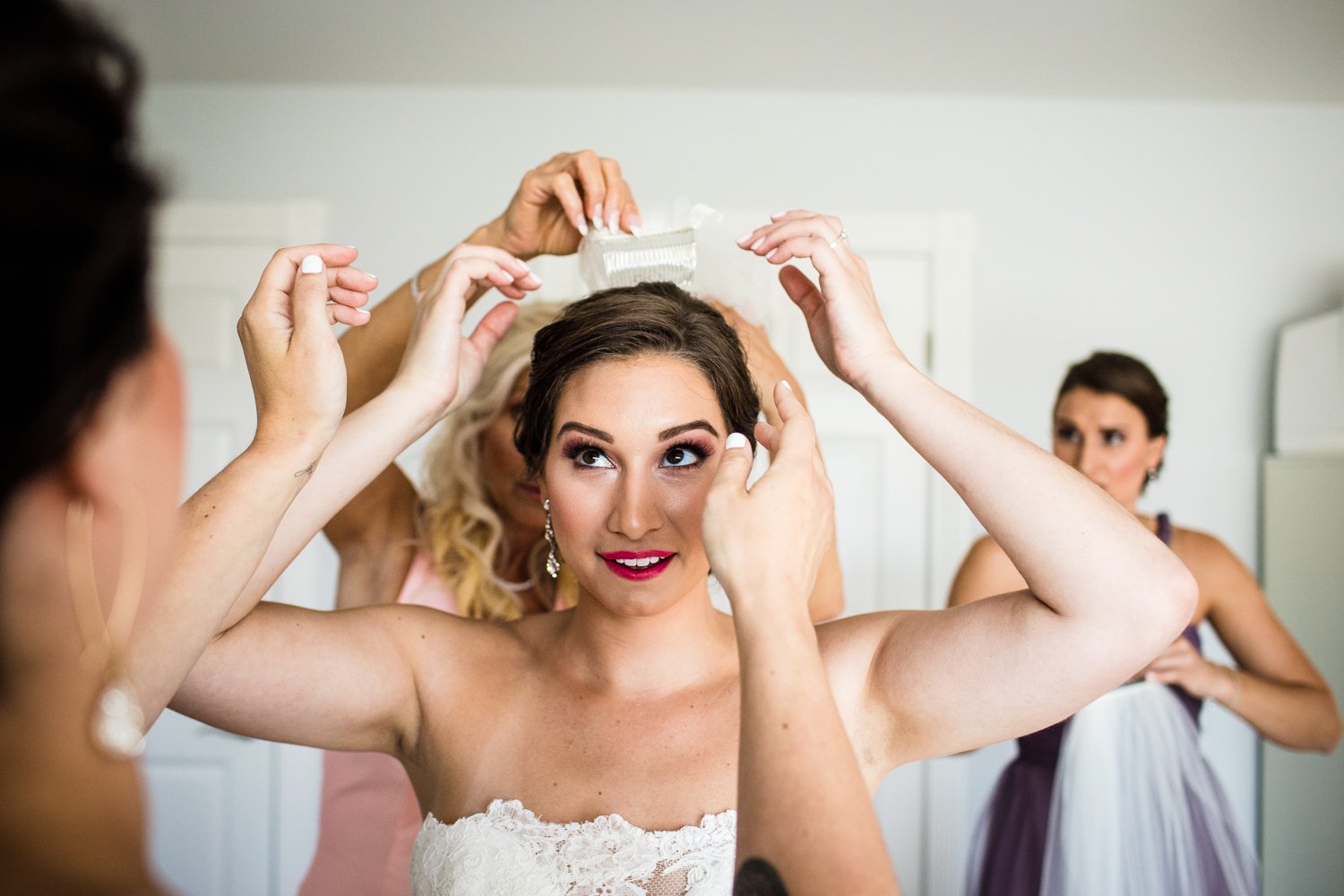 Bridesmaids help a bride put on her veil while getting ready together. 