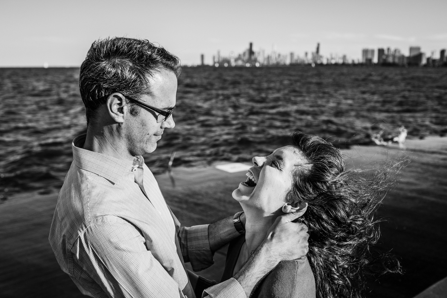 A couple laughs together in the wind with the Chicago skyline in the background during a Montrose Harbor engagement session. 