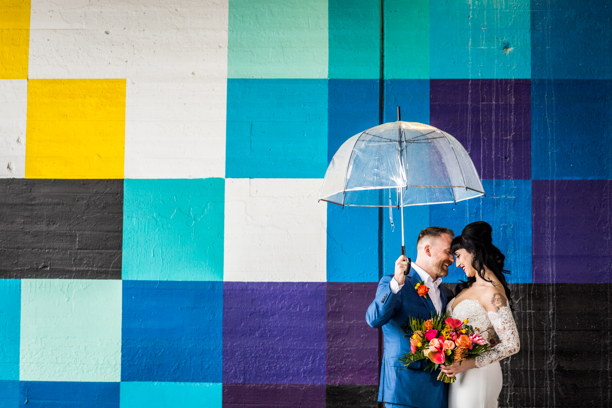 A couple stands under an umbrella in front of a Pantone wall before their Salvage One wedding. 