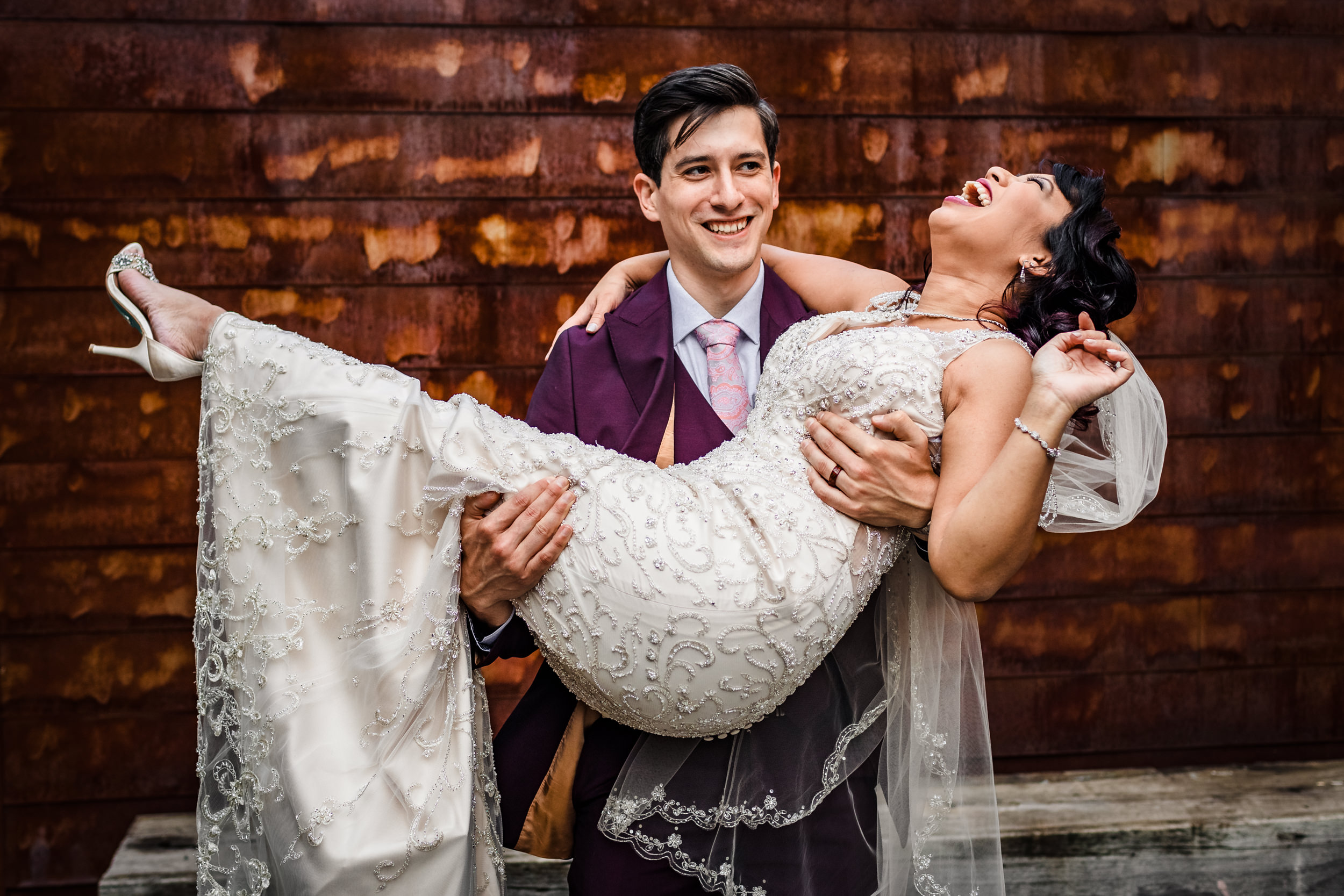 A groom carries his bride while laughing together after their wedding at The Joinery.